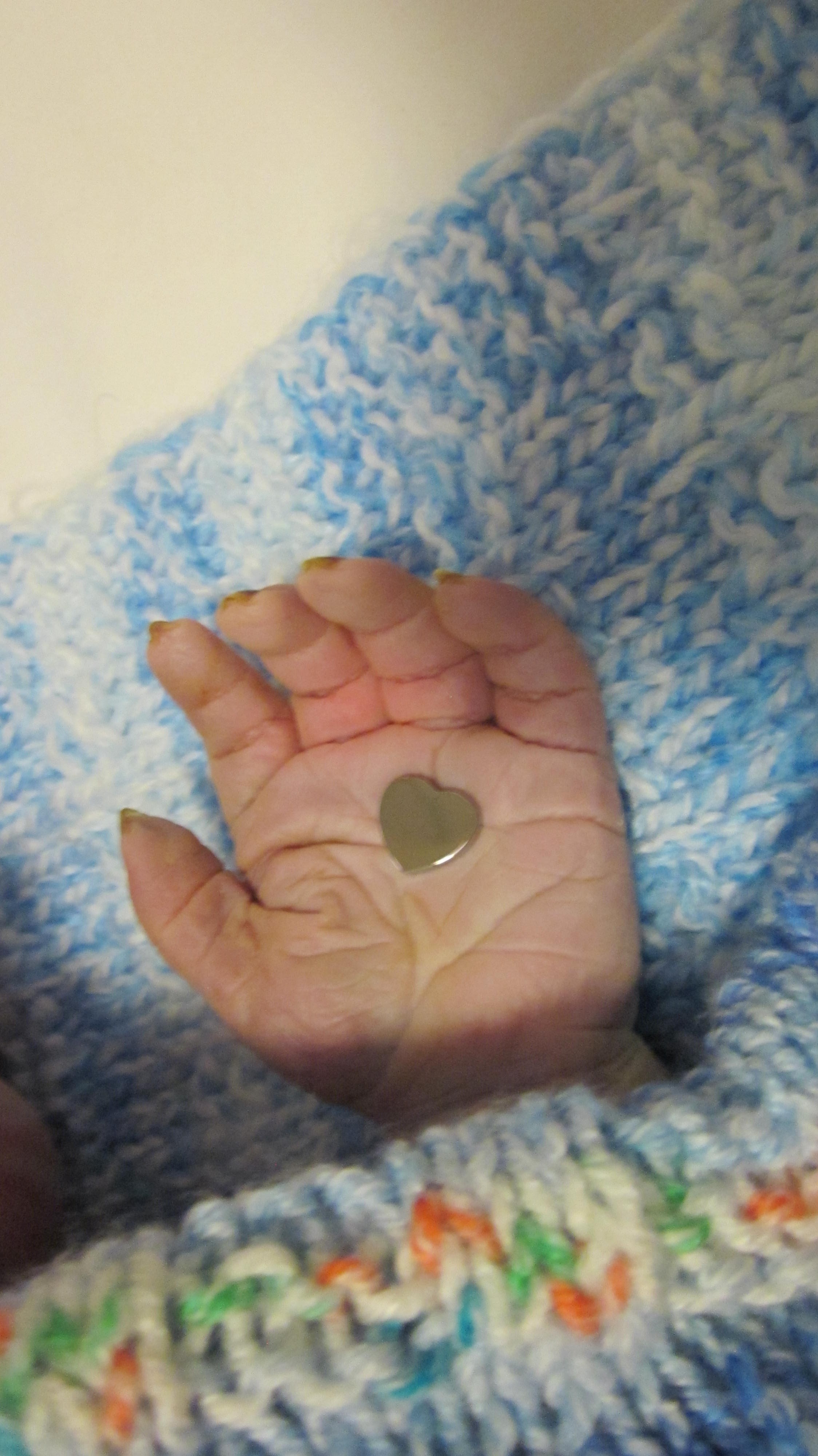 Baby hand with a silver heart laid in the palm on a white and blue knitted blanket