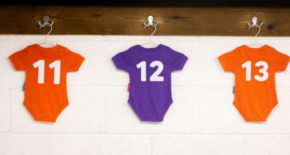 Three baby grows hang in a football dressing room. On the orange and purple baby grows are the numbers 11, 12 and 13