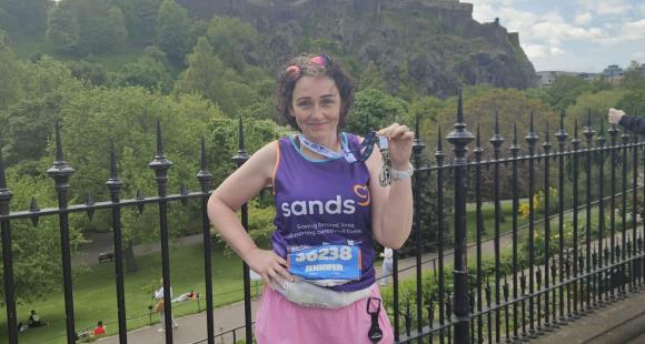A woman standing holding a medal up with her hand, and Edinburgh Castle behind her.