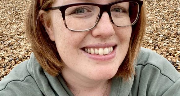 Lady with ginger hair and glasses smiling at the camera whilst sitting on a beach