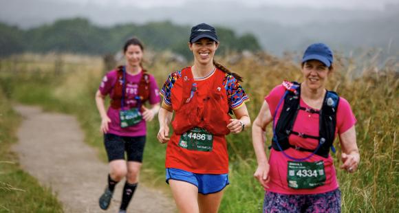 Three women running on a path with long grass in background