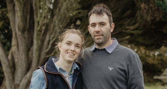 A lady with blonde hair tied up wearing a black jacket smiling at the camera with a man with dark hair with his arm around her wearing a grey jumper smiling at the camera with a background of trees.