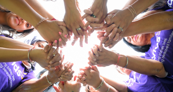 A group of women of different ethnicities hold hands in a circle. They are all wearing purple branded Sands tshirts