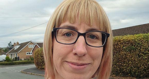 A lady with blonde hair and a fringe and black glasses smiling at the camera after her experience of missed miscarriage.