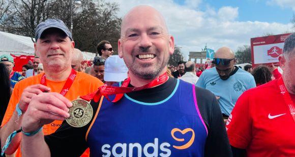 A man wearing a purple Sands running vest holding a gold medal up and smiling at the camera after running the London Marathon.
