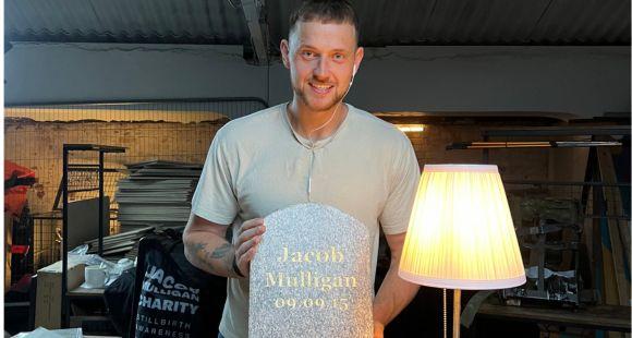 Man in a grey t-shirt smiling at the camera and holding a grey small headstone that reads Jacob Mulligan in gold letters 09.09.15 after his son was stillborn.