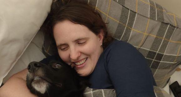 A lady in bed cuddling a black dog after talking about her miscarriages.