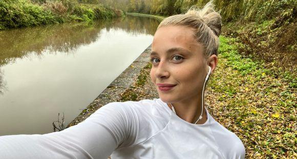 A lady with blonde hair in a top knotand a long white t-shirt smiling at the camera on a run with greenery in the background