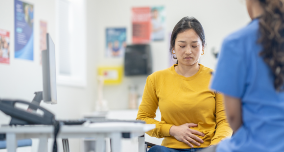 Woman holding her stomach looks down, whilst listening to a healthcare professional in blue scrubs