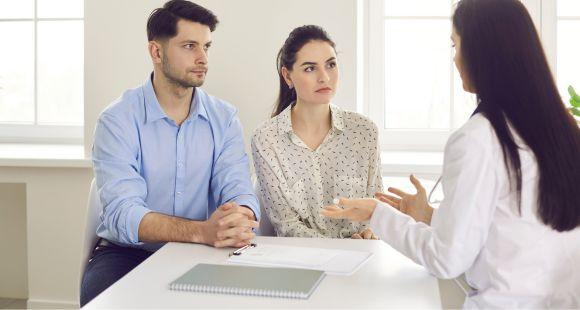 A white man withshort dark hair wearing a blue shirt and a white woman with long dark hair wearing a beige shirt sit at a desk facing a female healthcare worker with long dark hair wearing a white lab coat. She has her back to the camera and is gesturing with her hands. The man and woman look serious but calm.