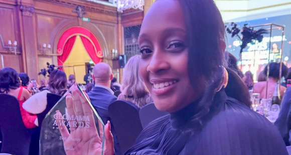 A woman dressed in black smiles while holding a clear “GloMama Awards – Best Charity” trophy at a formal awards ceremony, with guests seated behind her.