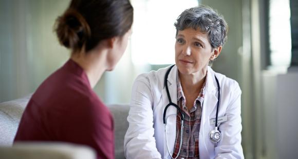A female healthcare worker with short grey hair wearing a white coat with a stethoscope around her neck looks with an expression of understanding toward a women with her back to the camera