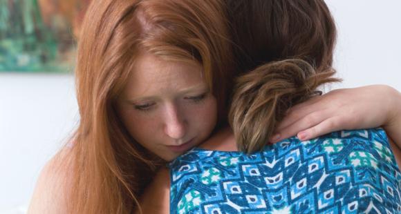 A woman with red hair embraces a woman with her back to the viewer, wearing a blue and white print dress