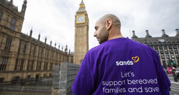 A male campaigner walks towards the houses of parliament in London. His purple Sands branded tshirt reads "Let's support bereaved parents and save babies' lives"