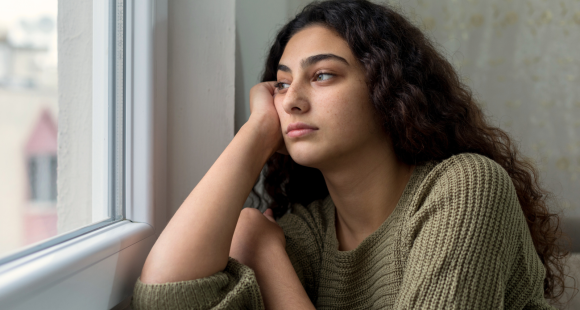 A young woman with long dark hair is looking out of a window with a sad expression