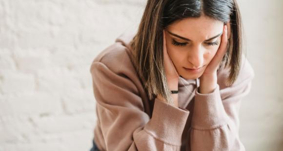 A young woman with bobbed brown hair wearing a beige sweater sits looking worried and holding her head in her hands 