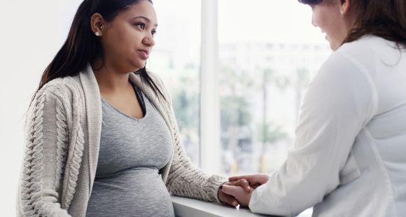 A white female healthcare worker wearing a white coat holds the hand of a pregnant Asian woman while speaking to her