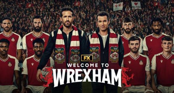 Show image of Rob McElhenny and Ryan Reynolds standing in front of Wrexham football team wearing club scarfs