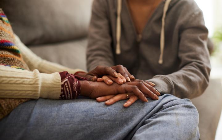Two people sat together on a sofa with hands on top of each other's to show support