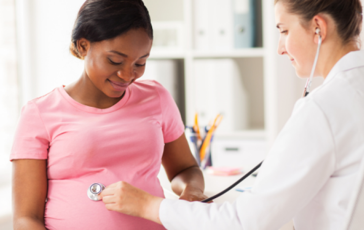 A female doctor in a white coat uses a stethoscope on the stomach of a pregnant Black woman wearing a pink top
