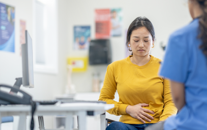 Woman holding her stomach looks down, whilst listening to a healthcare professional in blue scrubs
