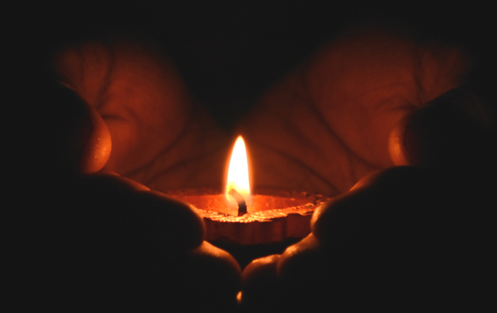 Two hands holding a diya candle, a close zoom in on the candle, very dark background 