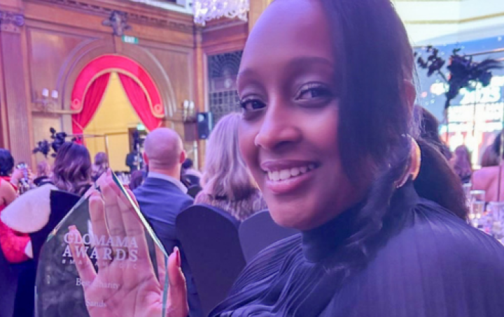 A woman dressed in black smiles while holding a clear “GloMama Awards – Best Charity” trophy at a formal awards ceremony, with guests seated behind her.