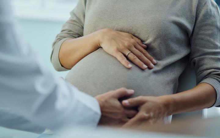 A pregnant woman wearing a beige jumper with her hand on her stomach faces a healthcare professional - you can not see either person's face