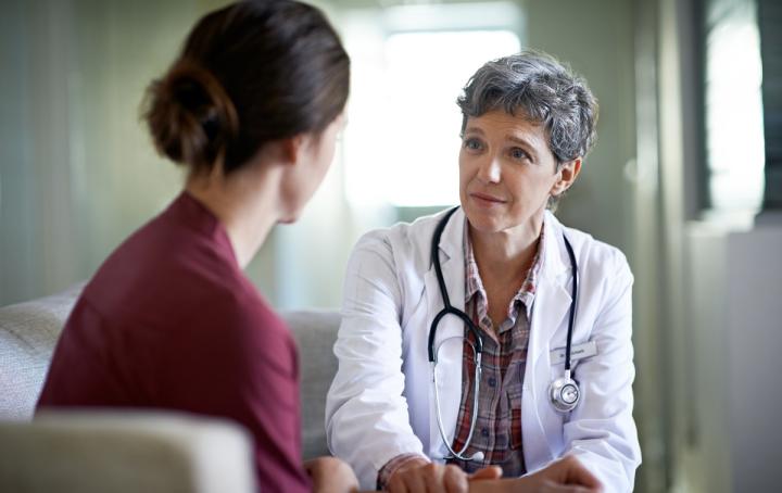 A female healthcare worker with short grey hair wearing a white coat with a stethoscope around her neck looks with an expression of understanding toward a women with her back to the camera