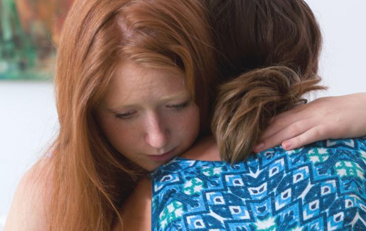 A woman with red hair embraces a woman with her back to the viewer, wearing a blue and white print dress