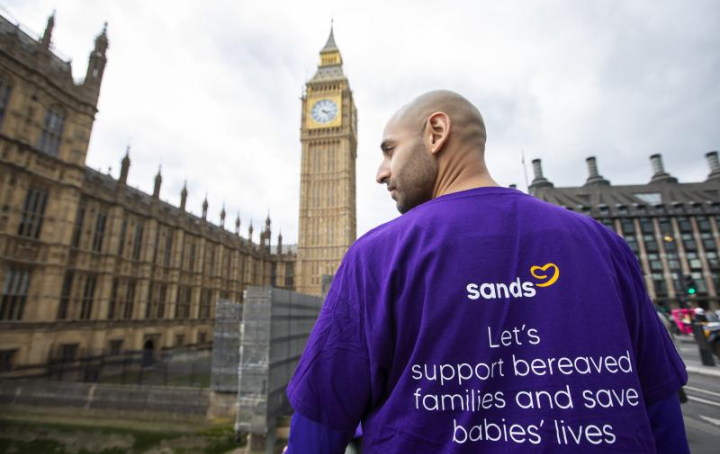 A male campaigner walks towards the houses of parliament in London. His purple Sands branded tshirt reads "Let's support bereaved parents and save babies' lives"