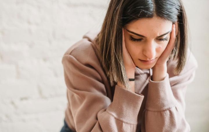 A young woman with bobbed brown hair wearing a beige sweater sits looking worried and holding her head in her hands 