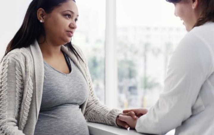 A white female healthcare worker wearing a white coat holds the hand of a pregnant Asian woman while speaking to her